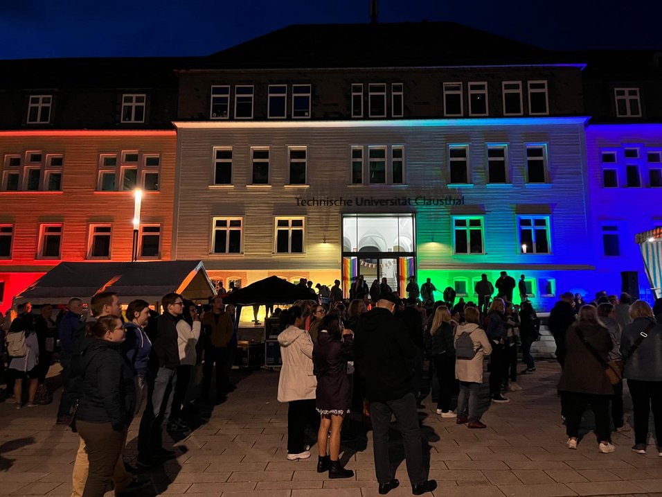 Hier ist das mit Regenbogenfarben angestrahlte TU Hauptgebäude mit einer Menschenmenge davor zu sehen.